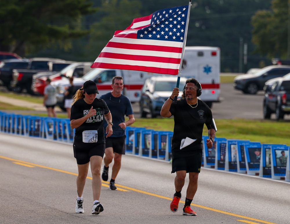 A group of runners participate in what appears to be a memorial run or patriotic race event. The lead runner carries a large American flag while running alongside others on a road lined with framed photographs or memorial placards. An ambulance and parked vehicles are visible in the background, suggesting an organized event with safety support.