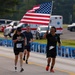 A group of runners participate in what appears to be a memorial run or patriotic race event. The lead runner carries a large American flag while running alongside others on a road lined with framed photographs or memorial placards. An ambulance and parked vehicles are visible in the background, suggesting an organized event with safety support.