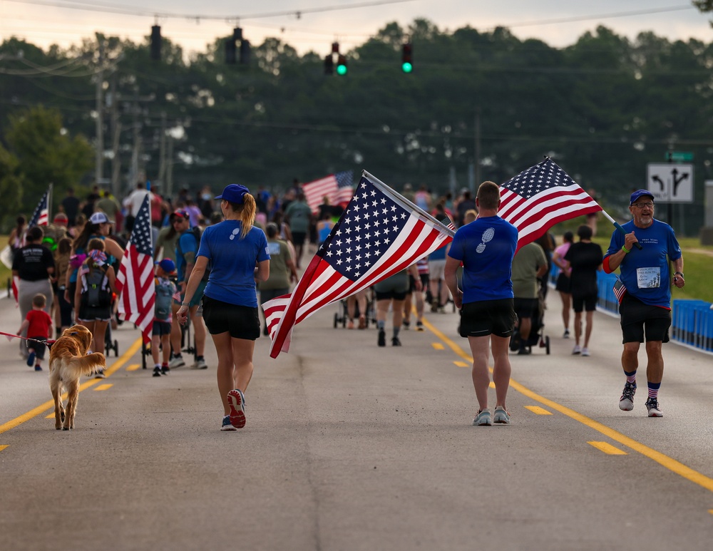 A large group of people are participating in what appears to be a patriotic walk or run event on a closed road, with multiple participants carrying large American flags. The event takes place at dusk or early morning, with participants wearing matching blue shirts and the crowd stretching far into the background. The scene suggests a community or military-affiliated patriotic event, possibly a 5K run or memorial march.