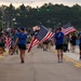 A large group of people are participating in what appears to be a patriotic walk or run event on a closed road, with multiple participants carrying large American flags. The event takes place at dusk or early morning, with participants wearing matching blue shirts and the crowd stretching far into the background. The scene suggests a community or military-affiliated patriotic event, possibly a 5K run or memorial march.
