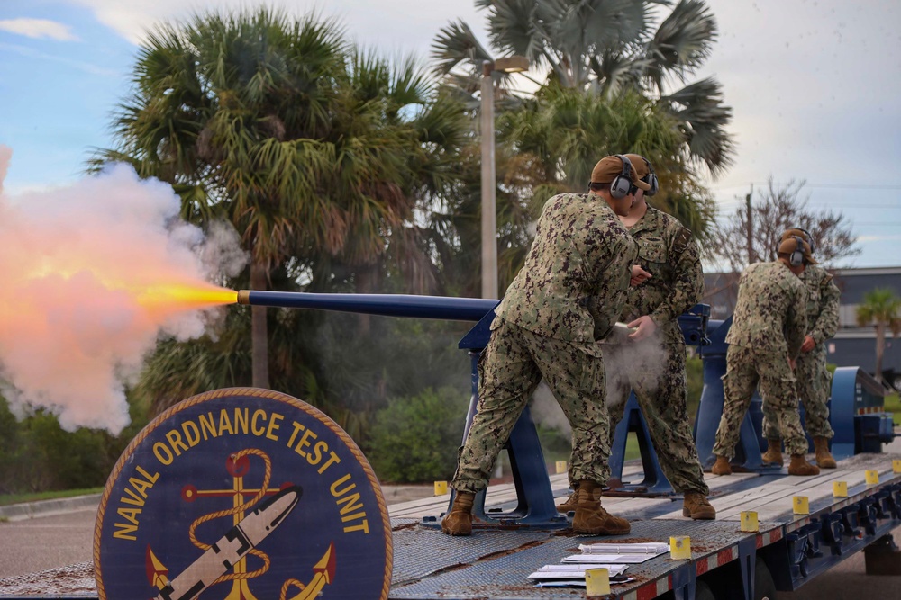 U.S. Navy personnel from the Naval Ordnance Test Unit are conducting a weapons test firing on a flatbed platform. A cannon or gun barrel is actively firing, producing a visible orange muzzle flash and white smoke plume to the left. At least three sailors in NWU Type III camouflage uniforms and hearing protection are present during the test.