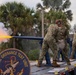 U.S. Navy personnel from the Naval Ordnance Test Unit are conducting a weapons test firing on a flatbed platform. A cannon or gun barrel is actively firing, producing a visible orange muzzle flash and white smoke plume to the left. At least three sailors in NWU Type III camouflage uniforms and hearing protection are present during the test.
