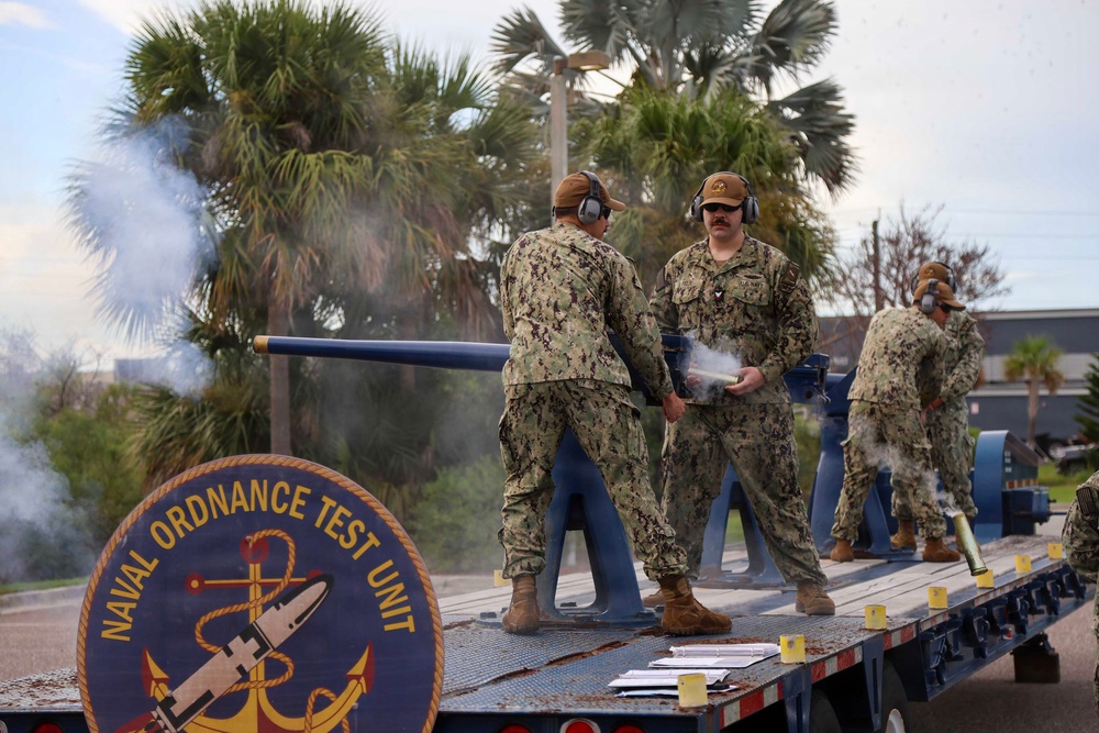 Personnel from the Naval Ordnance Test Unit are operating a deck gun or cannon mounted on a flatbed trailer, with visible smoke indicating a recent firing. At least three service members in U.S. Navy NWU Type III camouflage uniforms and hearing protection are actively handling the weapon and spent casings. The unit's official insignia patch is prominently displayed on the side of the trailer.