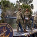 Personnel from the Naval Ordnance Test Unit are operating a deck gun or cannon mounted on a flatbed trailer, with visible smoke indicating a recent firing. At least three service members in U.S. Navy NWU Type III camouflage uniforms and hearing protection are actively handling the weapon and spent casings. The unit's official insignia patch is prominently displayed on the side of the trailer.