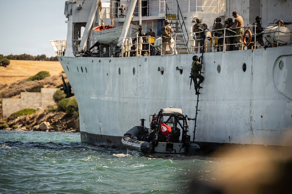 The image depicts a military maritime boarding operation, with armed personnel climbing the side of a large gray vessel using a rope ladder. A rigid inflatable boat (RIB) displaying a Tunisian flag is positioned alongside the ship, serving as the insertion platform. Multiple armed personnel in tactical gear are visible both on the deck of the ship and in the process of boarding.