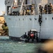 The image depicts a military maritime boarding operation, with armed personnel climbing the side of a large gray vessel using a rope ladder. A rigid inflatable boat (RIB) displaying a Tunisian flag is positioned alongside the ship, serving as the insertion platform. Multiple armed personnel in tactical gear are visible both on the deck of the ship and in the process of boarding.