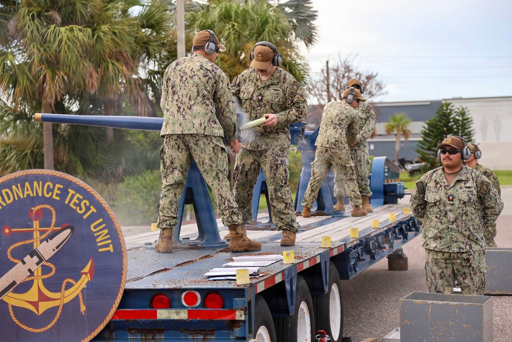 U.S. Navy personnel from an Ordnance Test Unit are conducting what appears to be a weapons or ordnance test on a flatbed trailer. Multiple sailors in NWU Type III camouflage uniforms and hearing protection are present, with visible smoke suggesting a recent discharge or test firing. The unit insignia on the trailer side reads 'Ordnance Test Unit' with a Navy anchor emblem.
