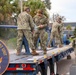 U.S. Navy personnel from an Ordnance Test Unit are conducting what appears to be a weapons or ordnance test on a flatbed trailer. Multiple sailors in NWU Type III camouflage uniforms and hearing protection are present, with visible smoke suggesting a recent discharge or test firing. The unit insignia on the trailer side reads 'Ordnance Test Unit' with a Navy anchor emblem.