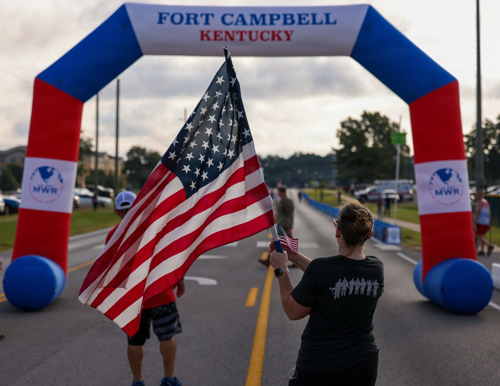 A woman in a black t-shirt waves a large American flag beneath an inflatable arch at Fort Campbell, Kentucky, during what appears to be a military community running event. The arch is decorated in red, white, and blue and bears the MWR (Morale, Welfare, and Recreation) logo. The scene suggests a patriotic race or fun run organized on the military installation.