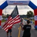 A woman in a black t-shirt waves a large American flag beneath an inflatable arch at Fort Campbell, Kentucky, during what appears to be a military community running event. The arch is decorated in red, white, and blue and bears the MWR (Morale, Welfare, and Recreation) logo. The scene suggests a patriotic race or fun run organized on the military installation.