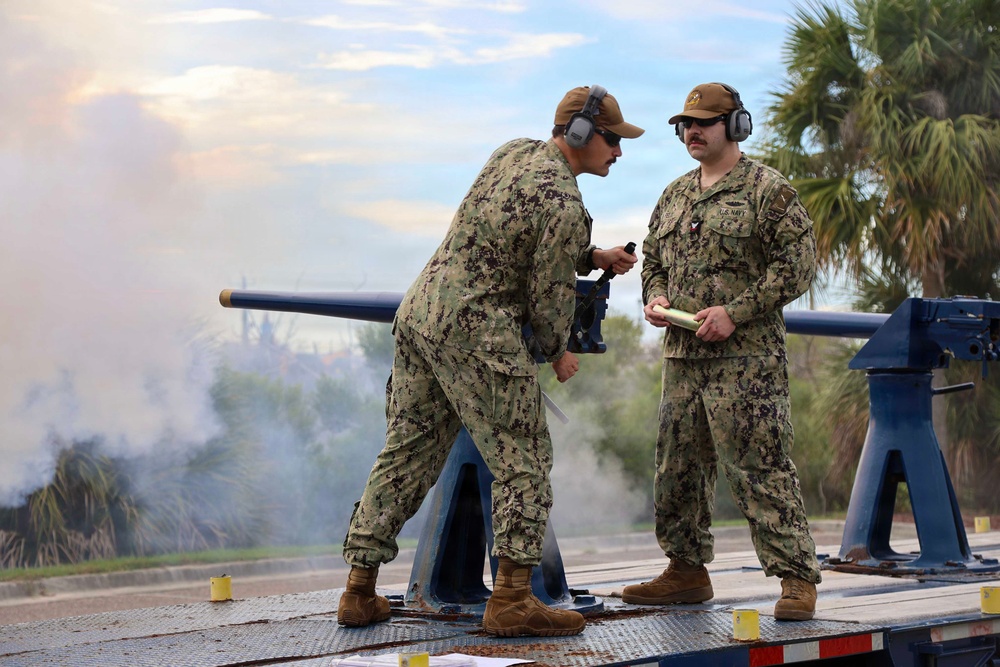 Two U.S. Navy personnel in NWU Type III camouflage uniforms are operating what appears to be a saluting cannon on a flatbed trailer platform. Smoke is visible in the background, suggesting the cannon has recently been fired. One sailor is leaning over the cannon while the other stands holding documents, both wearing hearing protection.
