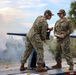 Two U.S. Navy personnel in NWU Type III camouflage uniforms are operating what appears to be a saluting cannon on a flatbed trailer platform. Smoke is visible in the background, suggesting the cannon has recently been fired. One sailor is leaning over the cannon while the other stands holding documents, both wearing hearing protection.