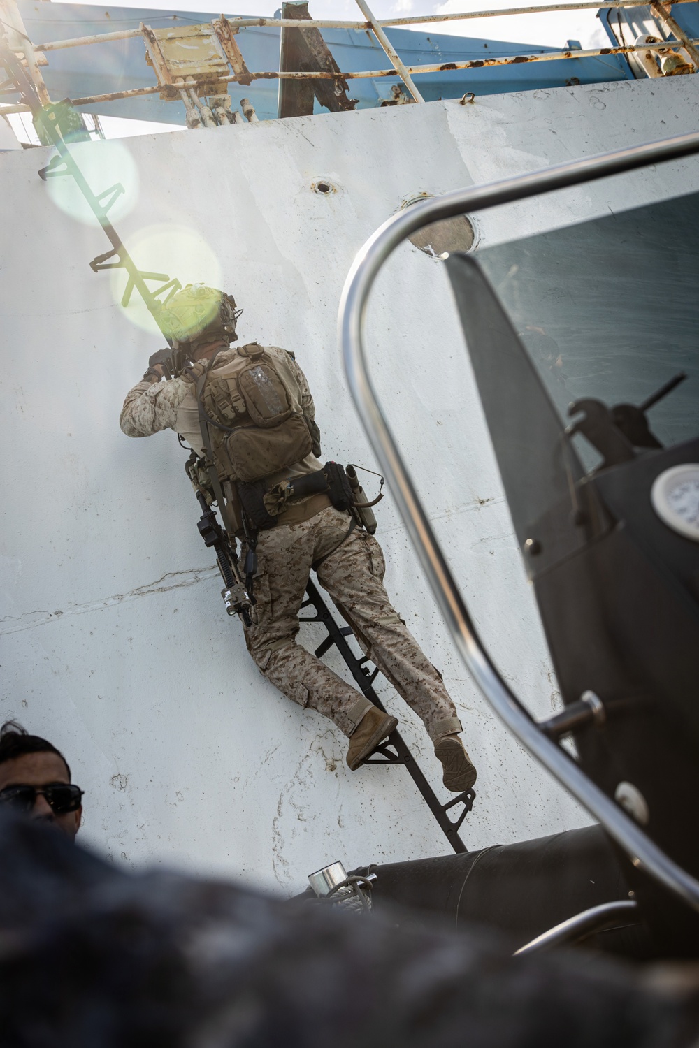 A military personnel in desert camouflage (MARPAT pattern) is climbing a ladder up the hull of a large vessel, consistent with a maritime boarding operation. The individual is fully equipped with tactical gear, body armor, a backpack, and a slung rifle. The scene appears to be taken from a smaller watercraft positioned alongside the larger ship.