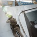 A military personnel in desert camouflage (MARPAT pattern) is climbing a ladder up the hull of a large vessel, consistent with a maritime boarding operation. The individual is fully equipped with tactical gear, body armor, a backpack, and a slung rifle. The scene appears to be taken from a smaller watercraft positioned alongside the larger ship.