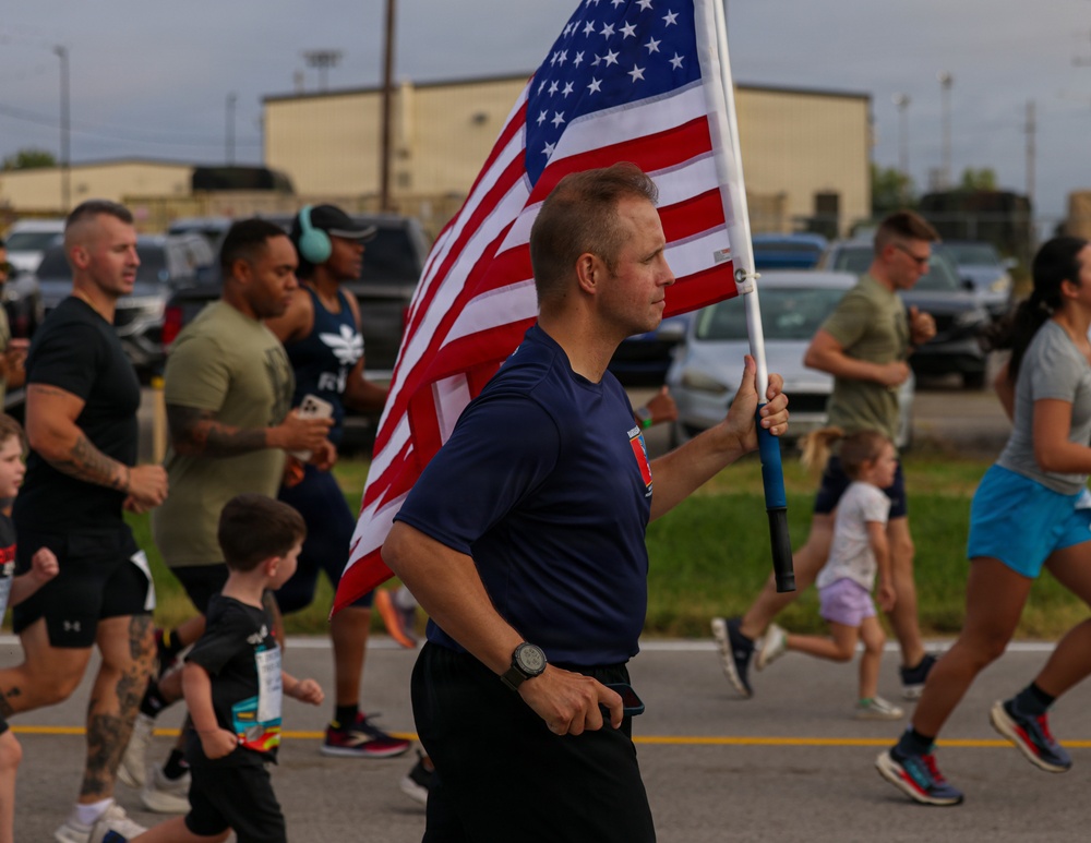 A group of adults and children are participating in an outdoor run or race event on a paved road. A central male figure in a navy blue shirt carries a large American flag while running. The event appears to be a patriotic or military-affiliated community run, likely held at or near a military installation based on the background buildings and participant attire.