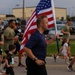 A group of adults and children are participating in an outdoor run or race event on a paved road. A central male figure in a navy blue shirt carries a large American flag while running. The event appears to be a patriotic or military-affiliated community run, likely held at or near a military installation based on the background buildings and participant attire.