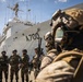 A group of military personnel in mixed uniforms and tactical gear are assembled on a dock in front of a large white naval vessel marked 'A700'. The scene appears to depict a joint military exercise or briefing between forces wearing different camouflage patterns, suggesting a multinational operation. The setting is a port environment under partly cloudy skies.