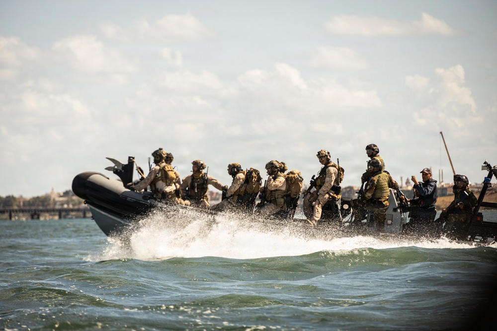 A group of heavily armed and equipped military personnel are traveling at high speed on a rigid inflatable boat (RIB) across open water. The operators are wearing tactical gear, helmets, and carrying weapons, suggesting a special operations or combat training exercise. A coastal cityscape or port infrastructure is faintly visible in the background.
