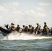 A group of heavily armed and equipped military personnel are traveling at high speed on a rigid inflatable boat (RIB) across open water. The operators are wearing tactical gear, helmets, and carrying weapons, suggesting a special operations or combat training exercise. A coastal cityscape or port infrastructure is faintly visible in the background.