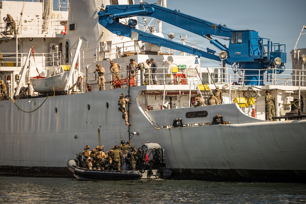 The image depicts a maritime tactical boarding operation, likely a military or law enforcement exercise, with armed personnel in combat gear ascending the hull of a large white vessel from a rigid inflatable boat (RIB). Multiple operatives are visible both on the small assault craft and already on the ship's deck, suggesting a coordinated ship-boarding drill. The vessel bears Arabic script on its hull and is equipped with a prominent blue crane structure.