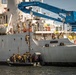 The image depicts a maritime tactical boarding operation, likely a military or law enforcement exercise, with armed personnel in combat gear ascending the hull of a large white vessel from a rigid inflatable boat (RIB). Multiple operatives are visible both on the small assault craft and already on the ship's deck, suggesting a coordinated ship-boarding drill. The vessel bears Arabic script on its hull and is equipped with a prominent blue crane structure.