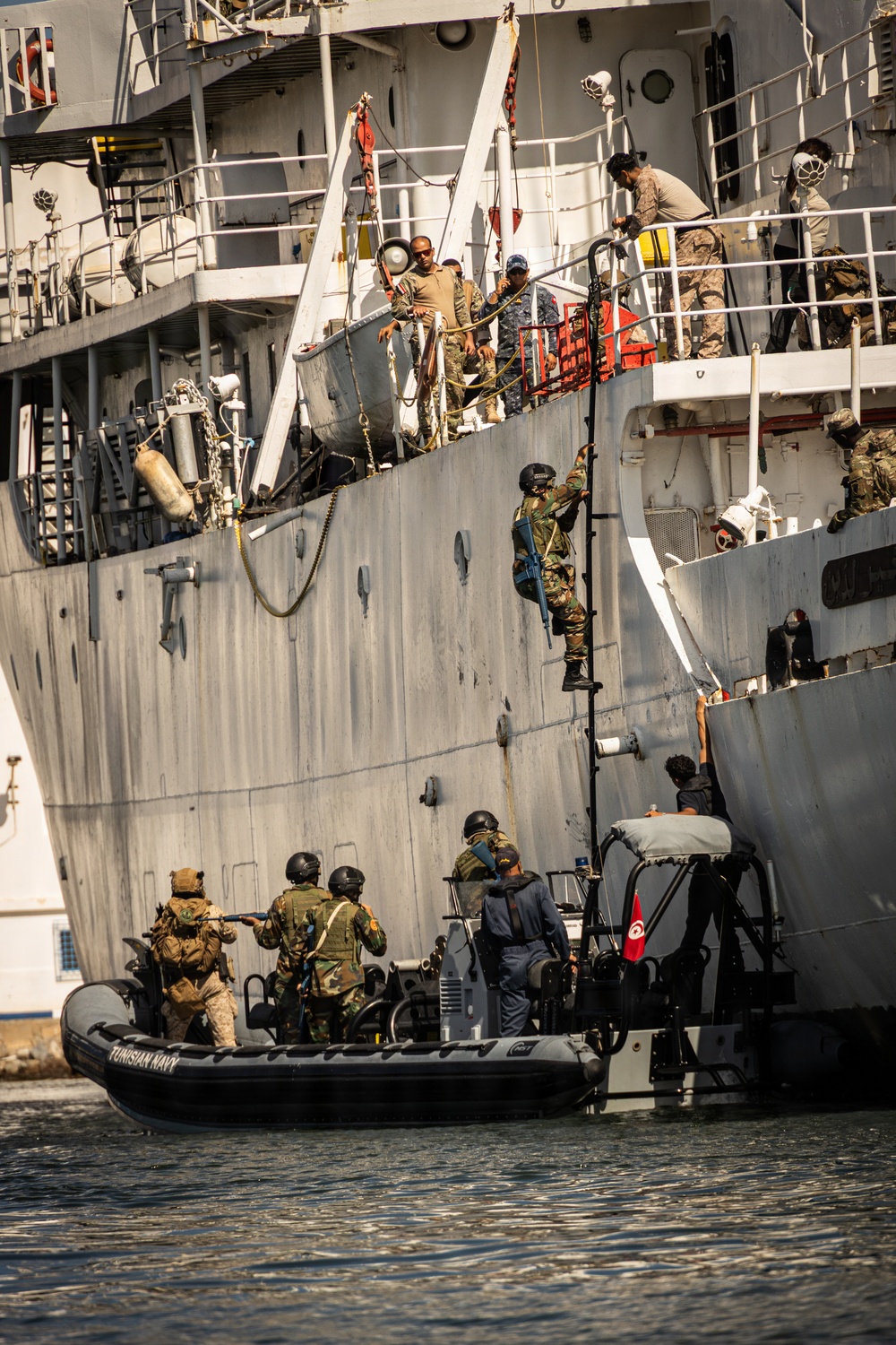 The image depicts a military maritime boarding exercise involving personnel from the Tunisian Navy, visible from the 'TUNISIAN NAVY' marking on the black rigid inflatable boat (RIB). Armed personnel in combat gear are climbing a rope ladder up the hull of a larger gray naval vessel while others remain on the RIB below. Personnel on the upper deck of the larger vessel are observing and assisting the boarding operation.