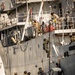 The image depicts a military maritime boarding exercise involving personnel from the Tunisian Navy, visible from the 'TUNISIAN NAVY' marking on the black rigid inflatable boat (RIB). Armed personnel in combat gear are climbing a rope ladder up the hull of a larger gray naval vessel while others remain on the RIB below. Personnel on the upper deck of the larger vessel are observing and assisting the boarding operation.