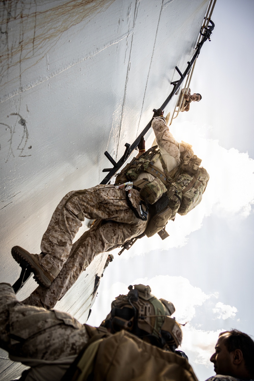 Military personnel in desert camouflage uniforms are conducting a vertical ship boarding or rappelling operation up the hull of a large vessel. The image is taken from a low angle looking upward, showing soldiers climbing a ladder or rope system along the white-painted ship hull. Multiple armed personnel are visible at various stages of the ascent, carrying full tactical gear and weapons.