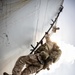 Military personnel in desert camouflage uniforms are conducting a vertical ship boarding or rappelling operation up the hull of a large vessel. The image is taken from a low angle looking upward, showing soldiers climbing a ladder or rope system along the white-painted ship hull. Multiple armed personnel are visible at various stages of the ascent, carrying full tactical gear and weapons.