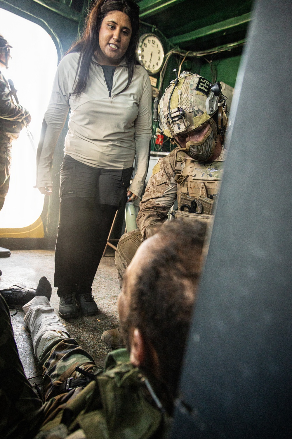The image shows what appears to be a military medical training or exercise scenario aboard a vessel or enclosed military facility. A fully equipped soldier in tactical gear and helmet is attending to a person lying on the floor, while a civilian woman in casual clothing observes. The setting includes green-painted walls, visible wiring, and a wall clock, suggesting a ship's interior or military installation.