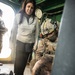 The image shows what appears to be a military medical training or exercise scenario aboard a vessel or enclosed military facility. A fully equipped soldier in tactical gear and helmet is attending to a person lying on the floor, while a civilian woman in casual clothing observes. The setting includes green-painted walls, visible wiring, and a wall clock, suggesting a ship's interior or military installation.