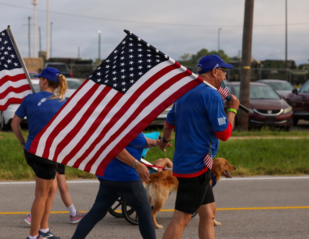 A group of people dressed in matching blue shirts and caps are marching along a road while carrying large and small American flags. A golden retriever dog is walking alongside the participants, and a stroller or cart is partially visible in the background. The scene appears to be a patriotic parade or commemorative walk event.