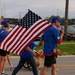 A group of people dressed in matching blue shirts and caps are marching along a road while carrying large and small American flags. A golden retriever dog is walking alongside the participants, and a stroller or cart is partially visible in the background. The scene appears to be a patriotic parade or commemorative walk event.