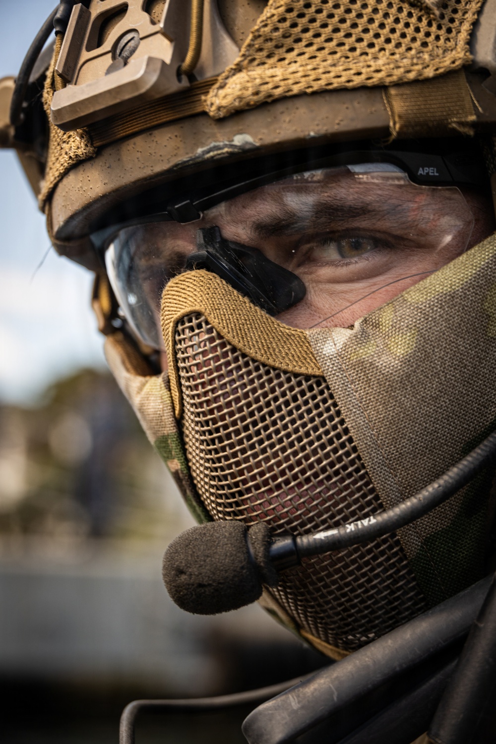 A close-up portrait of a military or tactical operator wearing full protective gear including a combat helmet, ballistic eyewear, mesh half-face mask, and a boom microphone. The individual's eyes are visible and appear focused, with face paint or camouflage markings visible on the skin. The equipment is in multicam/coyote tan coloration consistent with modern special operations gear.