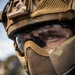 A close-up portrait of a military or tactical operator wearing full protective gear including a combat helmet, ballistic eyewear, mesh half-face mask, and a boom microphone. The individual's eyes are visible and appear focused, with face paint or camouflage markings visible on the skin. The equipment is in multicam/coyote tan coloration consistent with modern special operations gear.