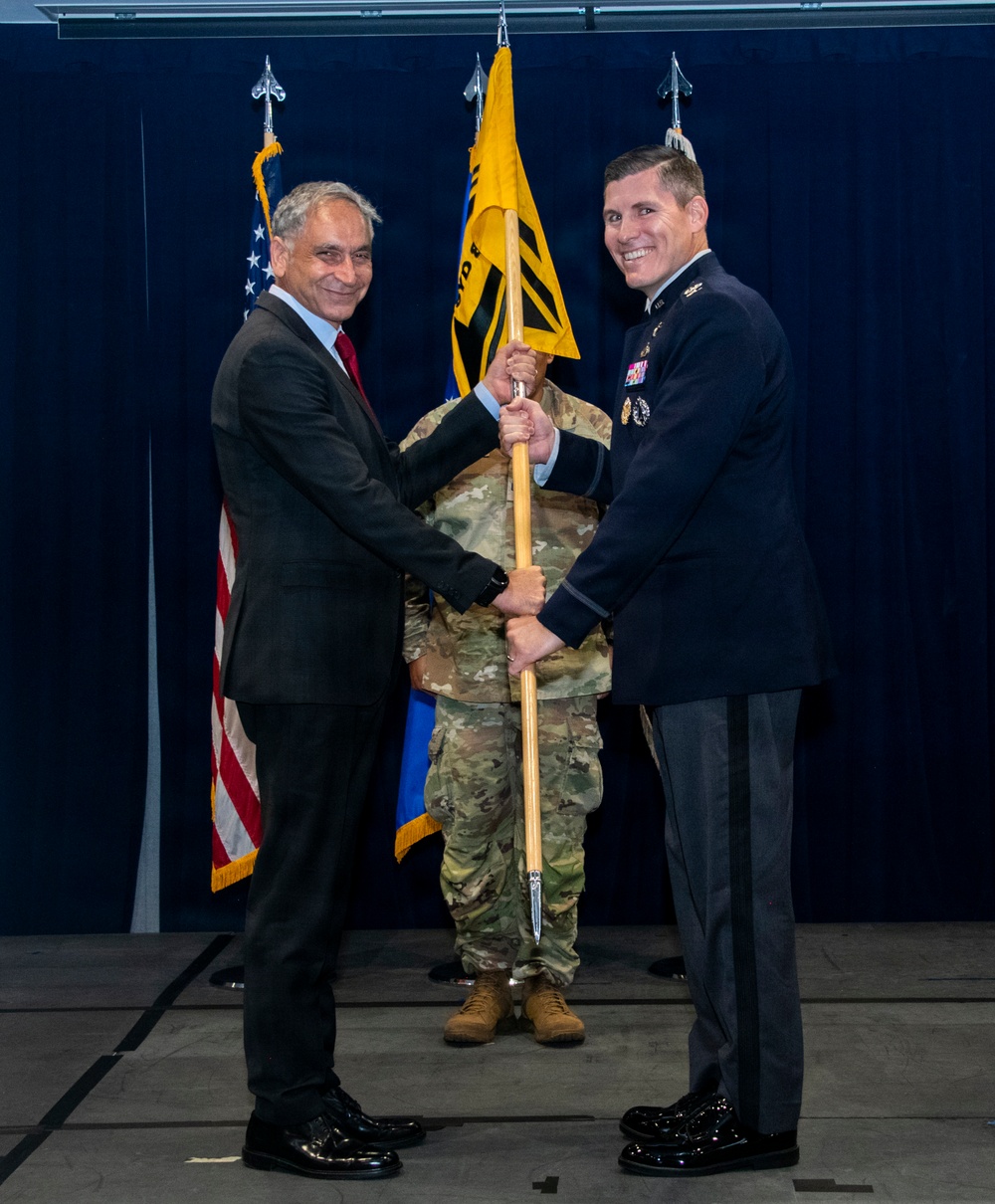 A formal military ceremony is taking place in which a civilian in a dark suit and a uniformed military officer in dress blues are jointly holding a guidon (unit flag) on a wooden staff. A third individual in camouflage uniform stands in the background holding additional flags. This appears to be a change of command or activation ceremony based on the guidon-passing tradition.