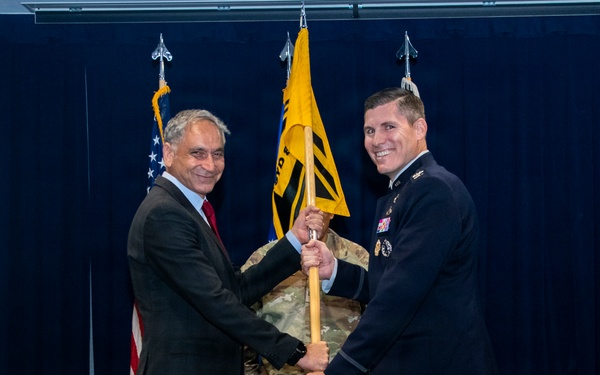 A formal military ceremony is taking place in which a civilian in a dark suit and a uniformed military officer in dress blues are jointly holding a guidon (unit flag) on a wooden staff. A third individual in camouflage uniform stands in the background holding additional flags. This appears to be a change of command or activation ceremony based on the guidon-passing tradition.