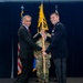 A formal military ceremony is taking place in which a civilian in a dark suit and a uniformed military officer in dress blues are jointly holding a guidon (unit flag) on a wooden staff. A third individual in camouflage uniform stands in the background holding additional flags. This appears to be a change of command or activation ceremony based on the guidon-passing tradition.