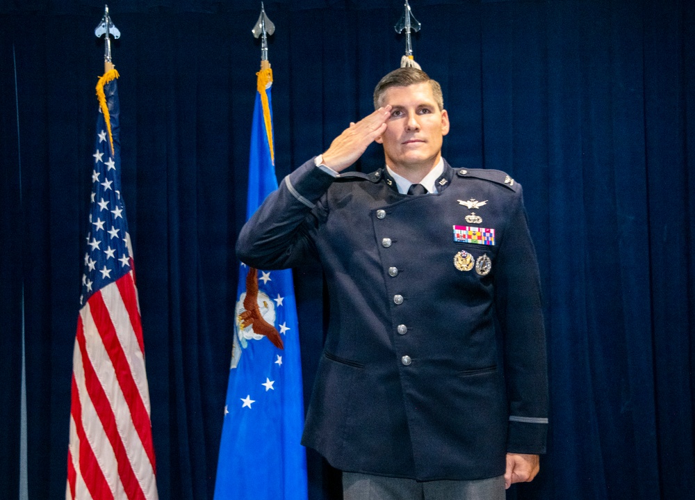A U.S. Air Force officer in dress uniform is rendering a salute during what appears to be a formal military ceremony. He is standing in front of the U.S. flag and the U.S. Air Force flag against a dark blue curtain backdrop. His uniform displays multiple medals, badges, and rank insignia consistent with a senior officer.