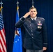 A U.S. Air Force officer in dress uniform is rendering a salute during what appears to be a formal military ceremony. He is standing in front of the U.S. flag and the U.S. Air Force flag against a dark blue curtain backdrop. His uniform displays multiple medals, badges, and rank insignia consistent with a senior officer.