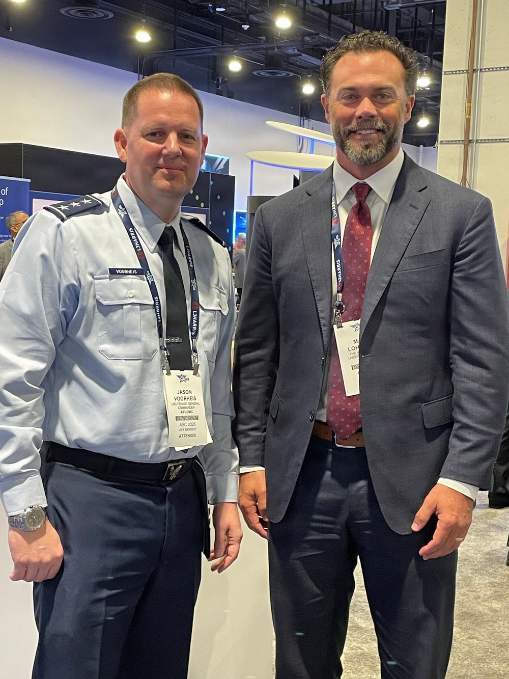 Two men pose for a photo at what appears to be a professional conference or expo, identifiable by the L3Harris-branded lanyards and name badges. The man on the left is in a U.S. Air Force uniform with three-star rank insignia, and his badge reads 'Jason Voorheis, Lieutenant General, Commander, AFLCMC, ASC 2025, AFA Member, Attendee.' The man on the right is in civilian business attire with a partially visible badge beginning with 'LOH.'