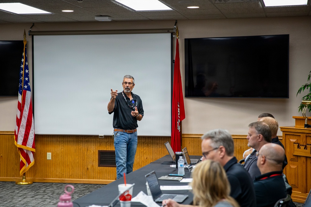 A man in a black shirt and jeans is presenting or speaking to a group of attendees seated at a conference table in a formal meeting room. The room contains a projection screen, a large wall-mounted TV, and both a U.S. flag and a red flag with insignia. Attendees appear to be listening attentively, with laptops and documents visible on the table.