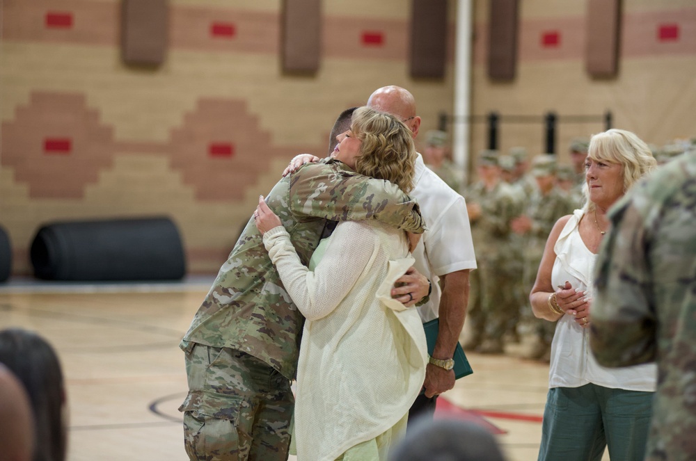 A military service member in camouflage uniform is embracing a civilian woman in what appears to be a gymnasium or indoor arena. Additional civilians and uniformed soldiers are visible in the background, suggesting this is a military ceremony such as a deployment send-off or homecoming event. The emotional embrace is the focal point of the image.