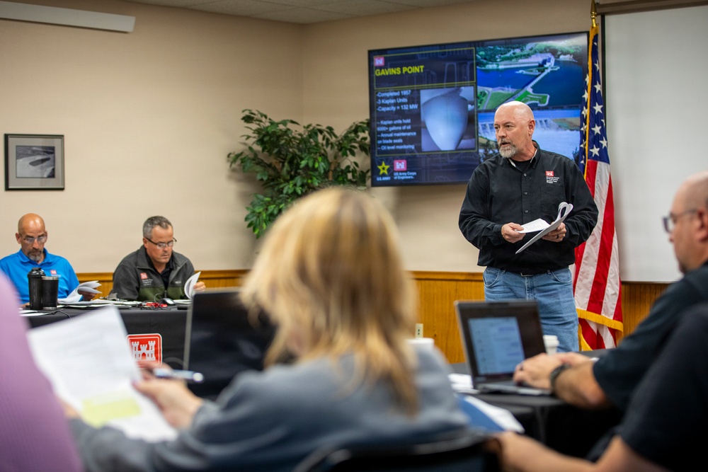 A briefing or meeting is taking place in a conference room where a man in a black shirt is presenting information about 'Gavins Point' dam/hydroelectric facility to a group of attendees. The presentation slide displayed on a large screen shows details about the Gavins Point project, including references to Kaplan units and 132 MW capacity, with the US Army Corps of Engineers logo visible. The setting appears to be an official government or military-affiliated meeting room.