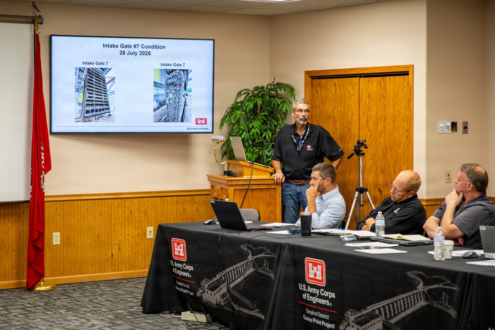 A U.S. Army Corps of Engineers briefing is taking place at the Gavins Point Project, Omaha District. A presenter stands at a podium delivering a presentation about 'Intake Gate #7 Condition' dated 26 July 2026, showing inspection photos on a large display screen. Three attendees are seated at a table draped with U.S. Army Corps of Engineers branded tablecloths.