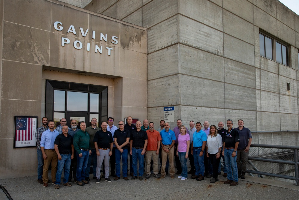 A large group of approximately 25 adults poses for a group photograph in front of the Gavins Point building, which appears to be a concrete government or infrastructure facility. The group consists predominantly of men with two visible women, dressed in casual to business-casual attire. A framed American flag display with '250 Years!' text is visible on the wall to the left of the entrance.