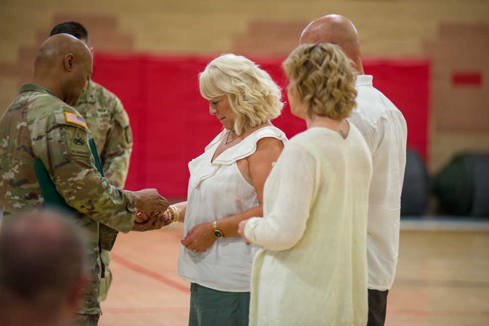 A U.S. Army soldier in OCP (Operational Camouflage Pattern) uniform is presenting or handing something to a blonde woman in a white top during what appears to be a military ceremony held in a gymnasium. The setting and body language suggest a formal recognition or award presentation event. Additional civilians, including a woman in a cream top and a bald man, stand nearby as participants or observers.