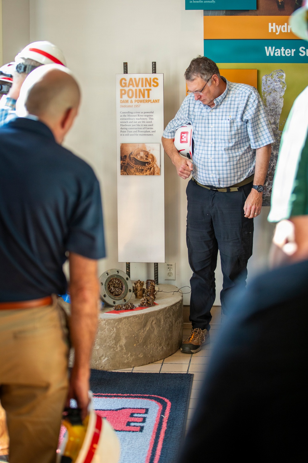 A group of visitors are gathered around an exhibit display at what appears to be a visitor center for the Gavins Point Dam &amp; Powerplant, dedicated in 1957. A man in a plaid shirt and glasses is leaning forward to closely examine the exhibit sign or artifacts on display. The exhibit features physical artifacts including what appear to be large bolts, nuts, and a flange, along with an informational panel.