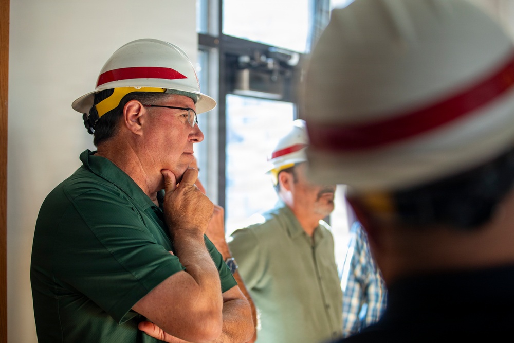 Three individuals wearing matching white hard hats with red stripes and yellow safety glasses are gathered in what appears to be a construction or industrial site interior. The foreground figure, wearing a green polo shirt, is in a contemplative pose with his hand on his chin and arms crossed, suggesting a briefing or inspection scenario. The matching personal protective equipment (PPE) across all visible individuals suggests they belong to the same organization or team.