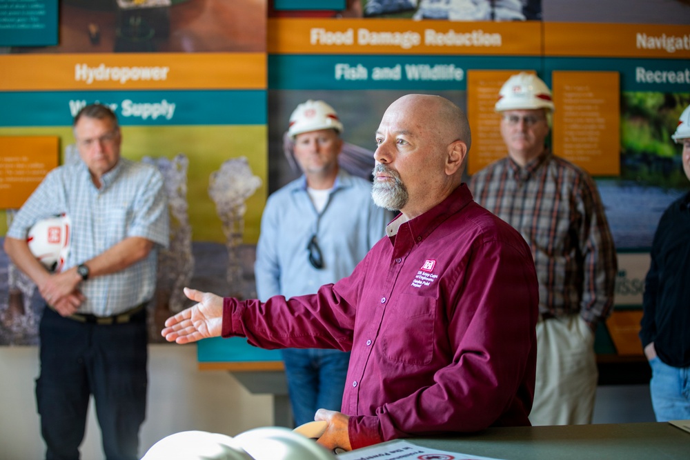 A man in a maroon US Army Corps of Engineers shirt is presenting or briefing a small group inside what appears to be a visitor center or educational facility. Behind him, informational display banners are visible covering topics such as 'Flood Damage Reduction,' 'Fish and Wildlife,' 'Hydropower,' and 'Water Supply.' Two individuals in the background are wearing white hard hats, suggesting a site visit or facility tour context.